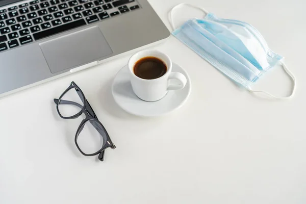 on the table is a medical mask and glasses, next is a coffee mug and a laptop