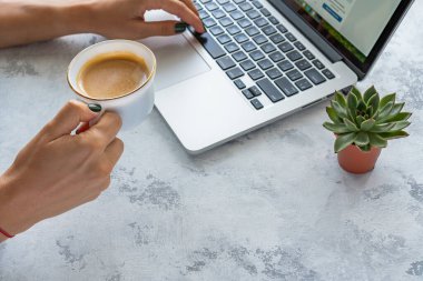 Bir kız elinde bir kupa kahve tutuyor, masanın yanında bir laptop var.