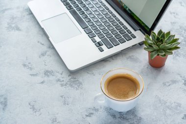 Masanın üzerinde bir dizüstü bilgisayar ve bir kupa kahve var. Yanında da saksıda bir çiçek var.