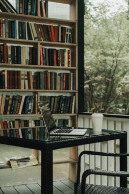 Sonbahar parkında üzerinde kahve olan bir laptop ve bir kupa var. Yanında bir kitaplık var.