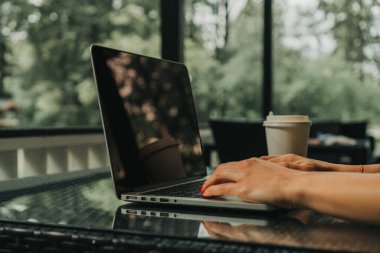 Kız bir kafede oturuyor ve dizüstü bilgisayarda klavye yazıyor. Yakınlarda bir fincan kahve var.