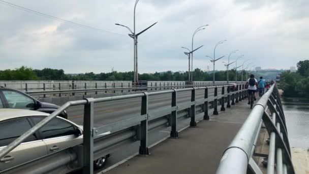 Ciclistas andam ao longo da ponte ao longo da auto-estrada. Tráfego de carros na rua. Dia nublado de verão. 4K. Kiev, Ucrânia . — Vídeo de Stock