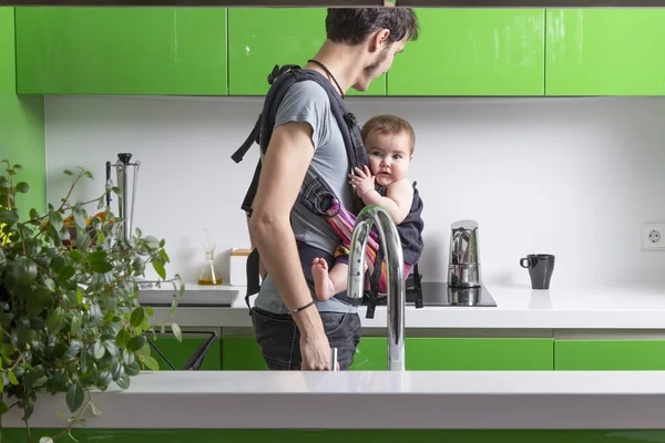 young father making coffee with his baby in carrier in green modern kitchen