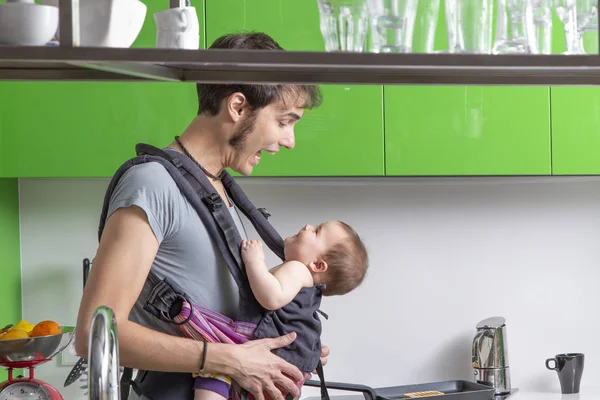 young father making coffee with his baby in carrier in green modern kitchen
