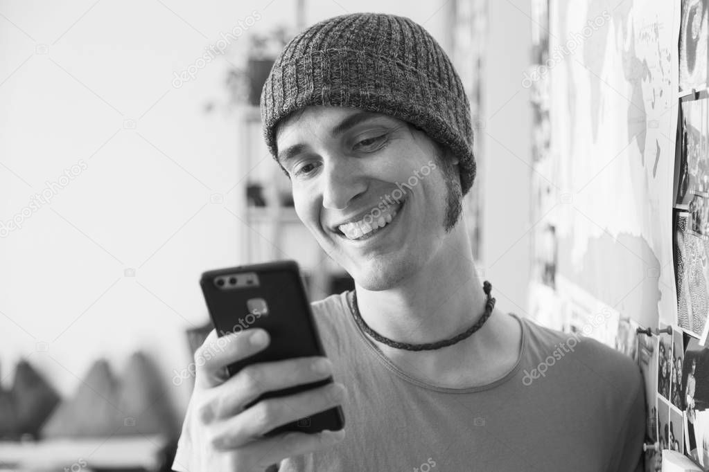 Black and white. Young man smiling with hat using smartphone at home. Whatsapp or tinder dating