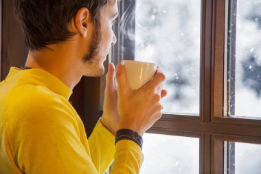 Kış sahne. Genç adam, kar pencereden bakarken sıcak bir kahve sarı giyinmiş