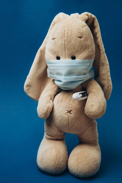 Un conejito de juguete lleva una máscara médica protectora con un termómetro debajo de su pata. — Foto de Stock