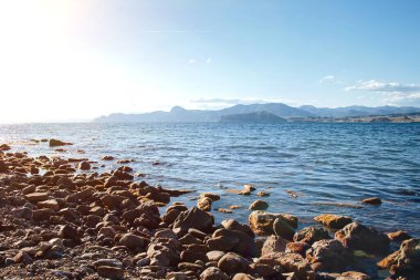 Mavi gökyüzü ve dağların arka planında deniz taşlı plaj.