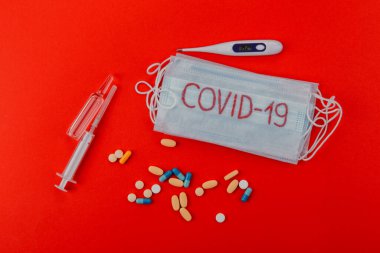 Üzerinde Covid-19 yazan koruyucu tıbbi maske, şırıngalar, termometre ampulleri ve kırmızı arka planda ilaçlar. Önlem olarak koruyucu maske.