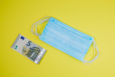 Tıbbi maske ve sarı arka planda para, dünya koronavirüs salgını ve ekonomik hasarlar. Corona virüsü konsepti.