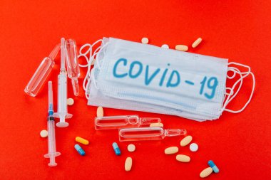 Üzerinde Covid-19 yazan koruyucu tıbbi maske kırmızı arka planda şırıngalar, ampuller ve ilaçlar. Yazı mavi ile yazılmış. Koronavirüsün yayılmasına karşı bir önlem olarak maske