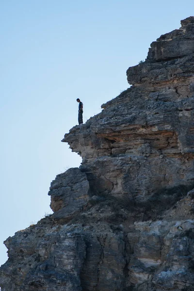 A man stands on the edge of a mountain against a blue sky. A tourist in the mountains stands on the rocks and looks down. Adventure, travel, active lifestyle.