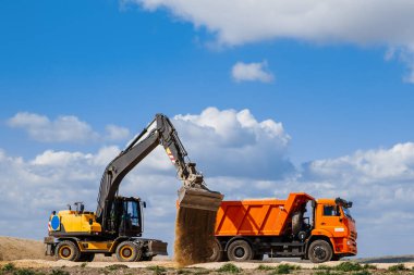 Sarı bir kepçe yükleyicisi bulutlu mavi gökyüzüne karşı bir yol inşa edilirken dünyayı bir kamyona yüklüyor..