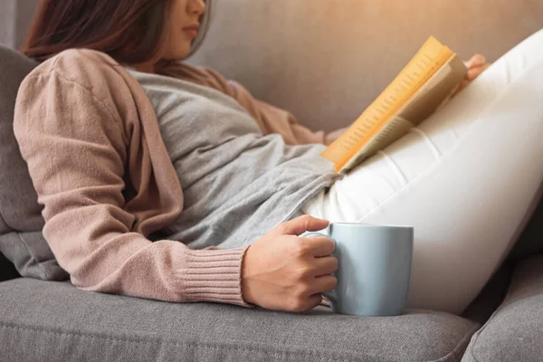 Elinde kahve fincanı tutan ve kanepede kitap okuyan genç bir kadın..