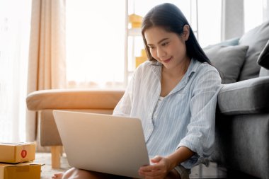 Küçük işletme sahibi kadın, çevrimiçi sipariş için müşteriye göndermek üzere ürünü kontrol etmek için dizüstü bilgisayar kullanıyor