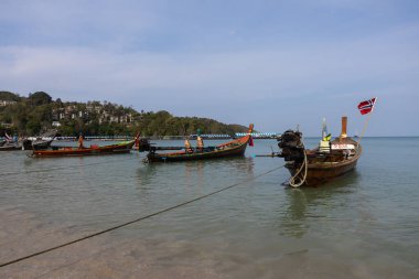 Phuket, Tayland - 06 Mart 2020: Tayland bayrağıyla Patong sahilinde geleneksel uzun kuyruklu tekneler. Arka planda ağaçlar ve evler var..