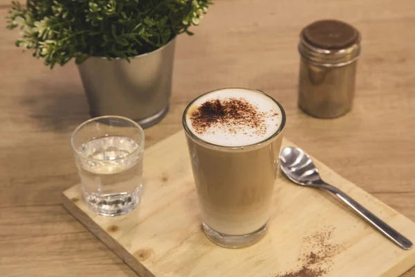 Glass of coffee with milk with glass of water and spoon on wooden board. Wooden background