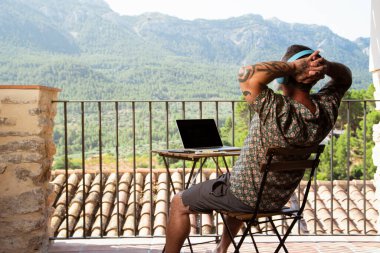 Yakışıklı bir adam evinin balkonunda dizüstü bilgisayarla çalışırken müzik dinliyor ve dağ manzarasının tadını çıkarıyor.