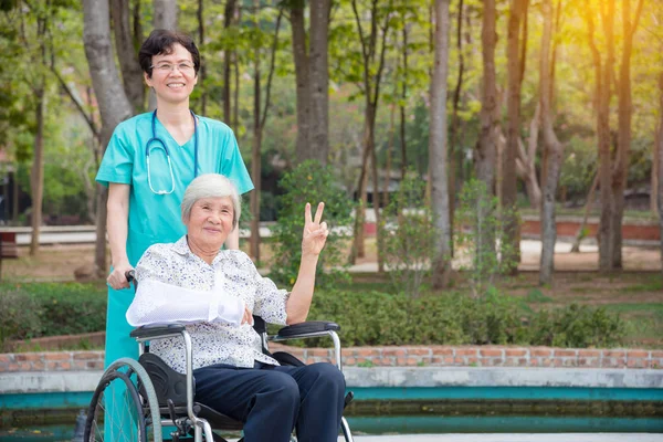 Feliz asiático senior mujer en silla de ruedas con enfermera en jardín —  Fotos de Stock