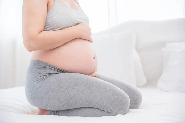 Gravid kvinna sitter på sängen och håller händerna på magen. — Stockfoto
