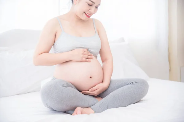 Gravid kvinna sitter på sängen och håller händerna på magen . — Stockfoto