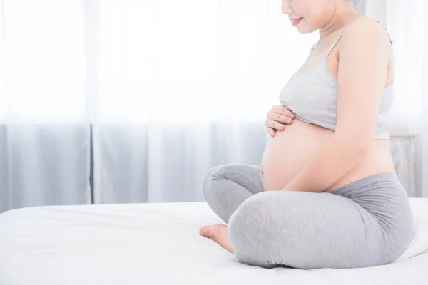 Беременная женщина сидит на кровати и держит руки на животе  . — стоковое фото