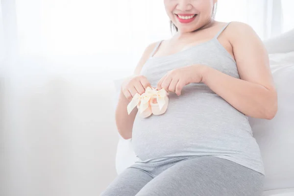 Gravid kvinna sitter på sängen och sätta lite baby skor på magen — Stockfoto