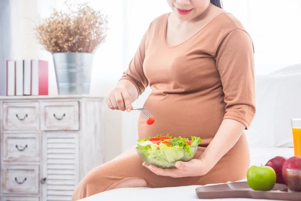 Donna incinta seduta sul letto, che mangia insalata a colazione . — Foto Stock