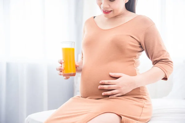 Donna incinta seduta sul letto, che tiene un bicchiere di succo d'arancia e sorride — Foto Stock
