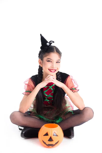 魔女の衣装で小さなアジアの女の子の完全な長さは座って、ハロウィーンのカボチャのバケツで笑顔 — ストック写真