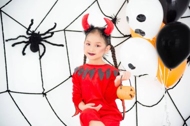 Kırmızı şeytan kıyafetli küçük Asyalı kız elinde balkabağı kovası ve cadılar bayramı süslemeli odasında gülümsüyor..