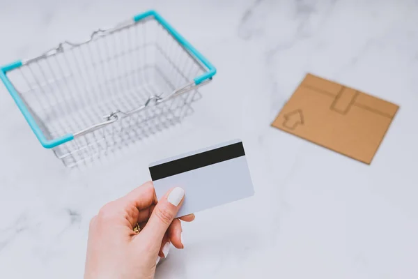 woman\'s hand holding payment card next to empty shopping basket and mini cardboard delivery parcel, concept of shopping online