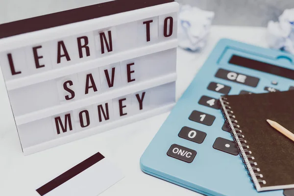 lightbox with Learn to save money message next to payment card calculator and stationery on marble desk, concept of learning to spend less and refrain from excessive shopping
