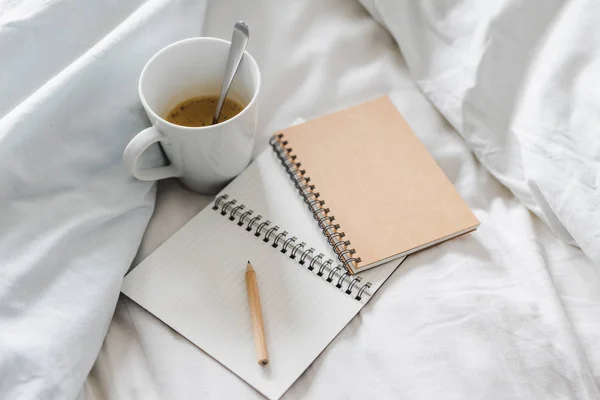 empty notepads with copyspace and pencil on bed with mug of coffee next to it, concept of starting the morning and writing a schedule or to do list