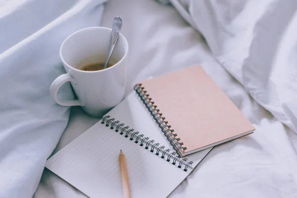empty notepads with copyspace and pencil on bed with mug of coffee next to it, concept of starting the morning and writing a schedule or to do list