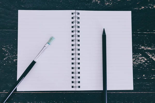 Notepad on black wooden desk with brush and pencil side by side — Stock Photo, Image