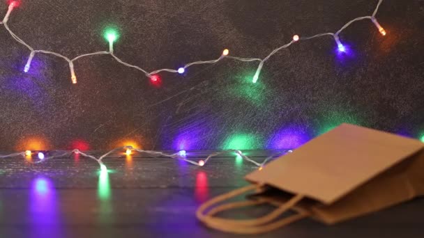 お祝いの季節のショッピングコンセプト 手は背景に多色のクリスマスストリングライトとバッグの隣に買い物かごを配置 — ストック動画
