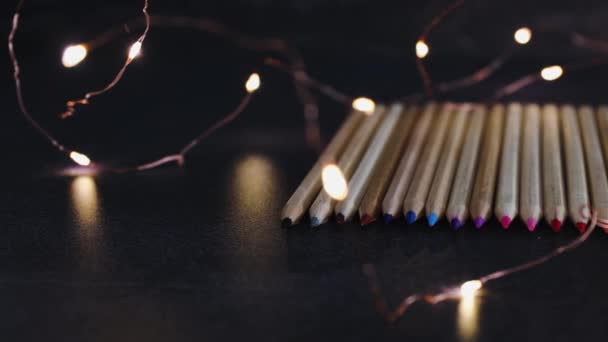 色付きの鉛筆は 妖精のライトに囲まれた暗い机の上に並び カメラが左右に水平にパンします — ストック動画