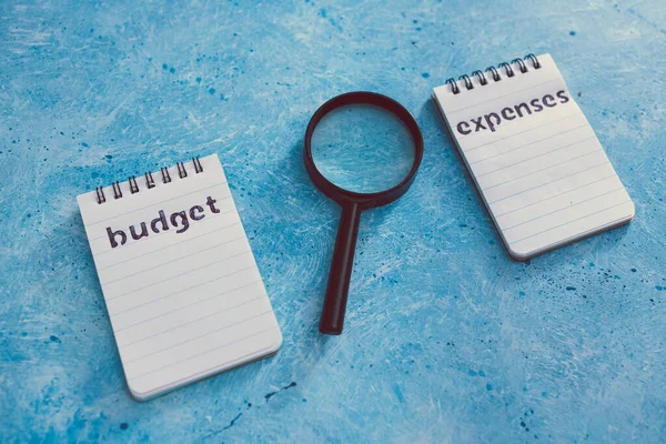 managing money and finance concept, budget and expenses notepads side by side on blue desk with magnifying glass