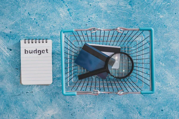 managing money and finance concept, budget notepad with shopping basket magnifying glass and credit cards on blue desk