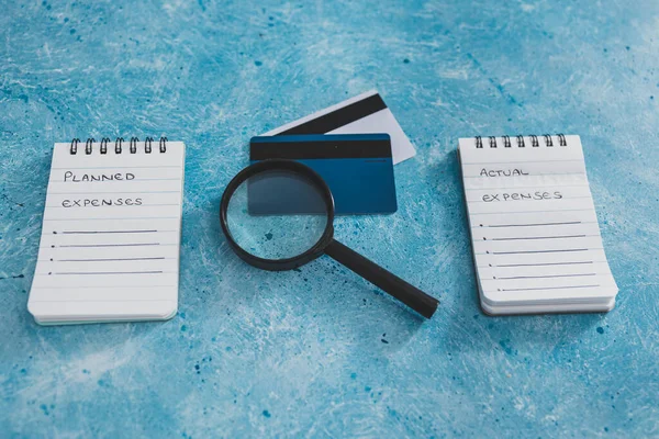 managing money and finance concept, planned expenses vs actual expenses notepads side by side with credit cards and magnifying glass on blue desk