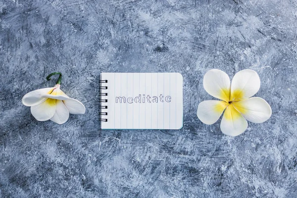 self-care and mental health concept, notepad with text Meditate next to tropical flowers on grey concrete surface