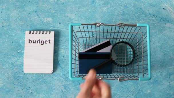 お金や金融の概念を管理し 予算のノートパッドを買い物かごに入れ 手で虫眼鏡やクレジットカードを青い机の上につかむ — ストック動画