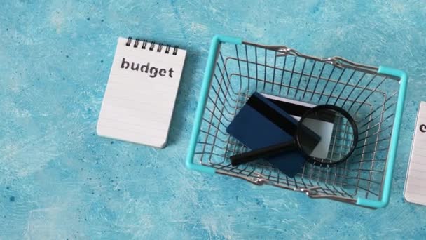 管理货币和财务概念 预算和支出记事本 用购物篮放大镜和蓝色桌子上的信用卡和相机套餐 — 图库视频影像