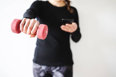 Evde fitness ve internet koçluğu konsepti, spor kıyafetleri içindeki kadının elinde dambıl ve akıllı telefon tutarak spor videosu ya da internetteki rutini takip etmesi.
