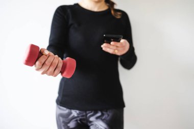 Evde fitness ve internet koçluğu konsepti, spor kıyafetleri içindeki kadının elinde dambıl ve akıllı telefon tutarak spor videosu ya da internetteki rutini takip etmesi.