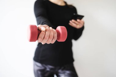 Evde fitness ve internet koçluğu konsepti, spor kıyafetleri içindeki kadının elinde dambıl ve akıllı telefon tutarak spor videosu ya da internetteki rutini takip etmesi.
