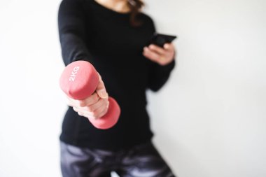 Evde fitness ve internet koçluğu konsepti, spor kıyafetleri içindeki kadının elinde dambıl ve akıllı telefon tutarak spor videosu ya da internetteki rutini takip etmesi.