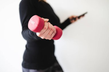 Evde fitness ve internet koçluğu konsepti, spor kıyafetleri içindeki kadının elinde dambıl ve akıllı telefon tutarak spor videosu ya da internetteki rutini takip etmesi.
