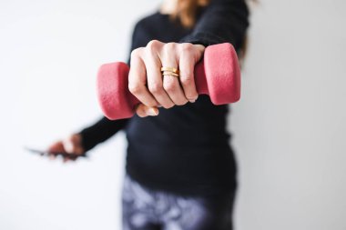 Evde fitness ve internet koçluğu konsepti, spor kıyafetleri içindeki kadının elinde dambıl ve akıllı telefon tutarak spor videosu ya da internetteki rutini takip etmesi.